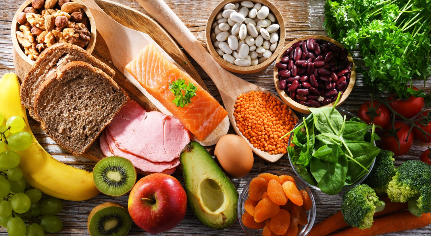 A vibrant flat lay overhead shot of a variety of fresh, healthy foods arranged on a wooden surface, including salmon, sliced ham, an avocado, nuts, eggs, and assorted fruits and vegetables.